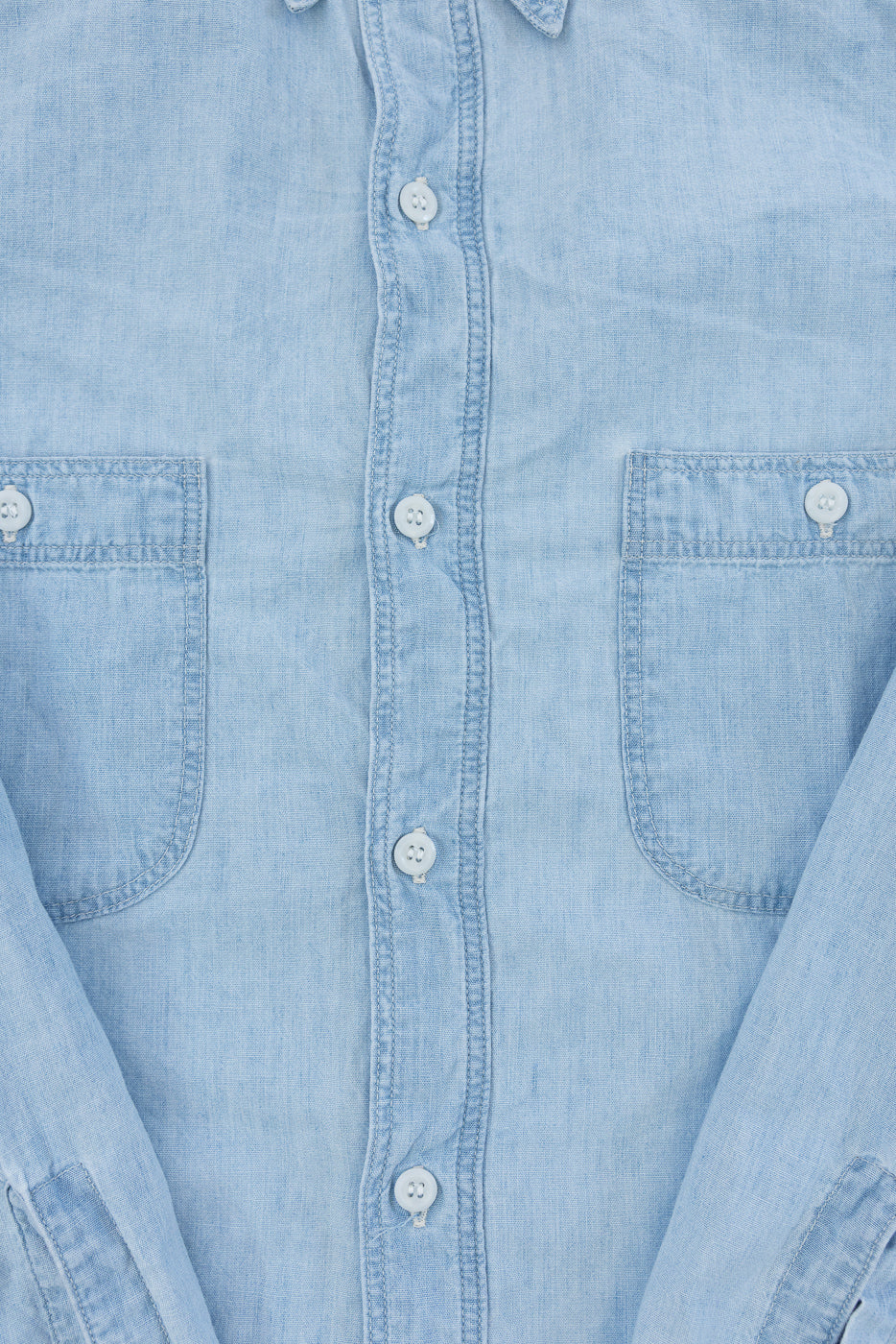 Close-up of front chest area of light blue chambray work shirt, showing buttons, two front pockets, and fabric texture.