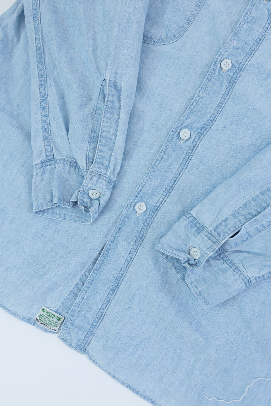 Close-up of front of light blue chambray work shirt showing buttons, cuffs, and hem tag.