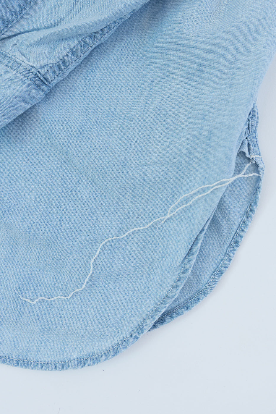 Close-up of chambray shirt hem, front bottom edge detail, showing fabric texture and loose thread.