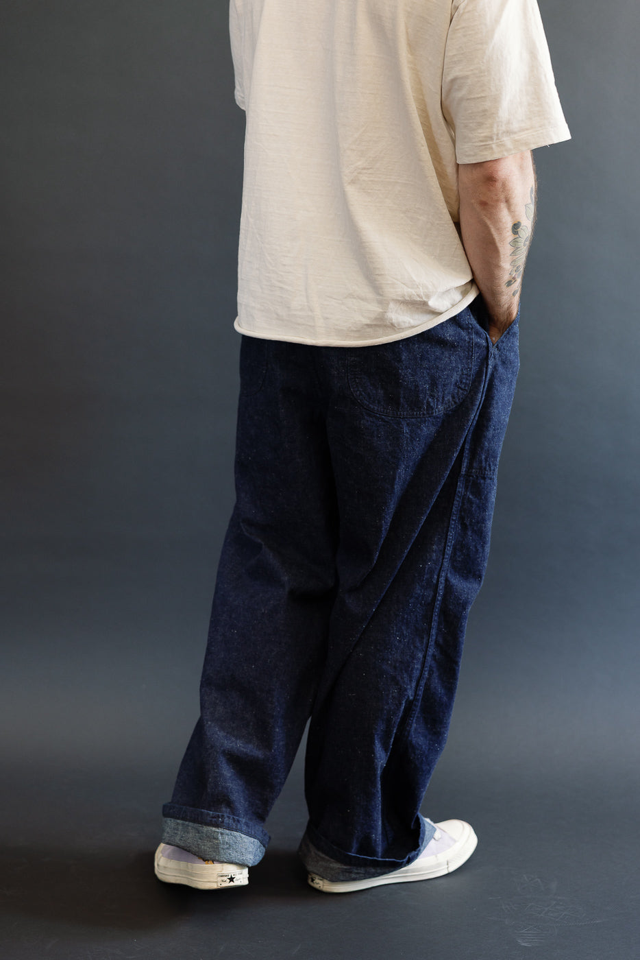 Full body rear view of male model 5'9, 165lbs, athletic build wearing indigo wide-leg selvedge denim utility pants with visible side seams and large back pockets.