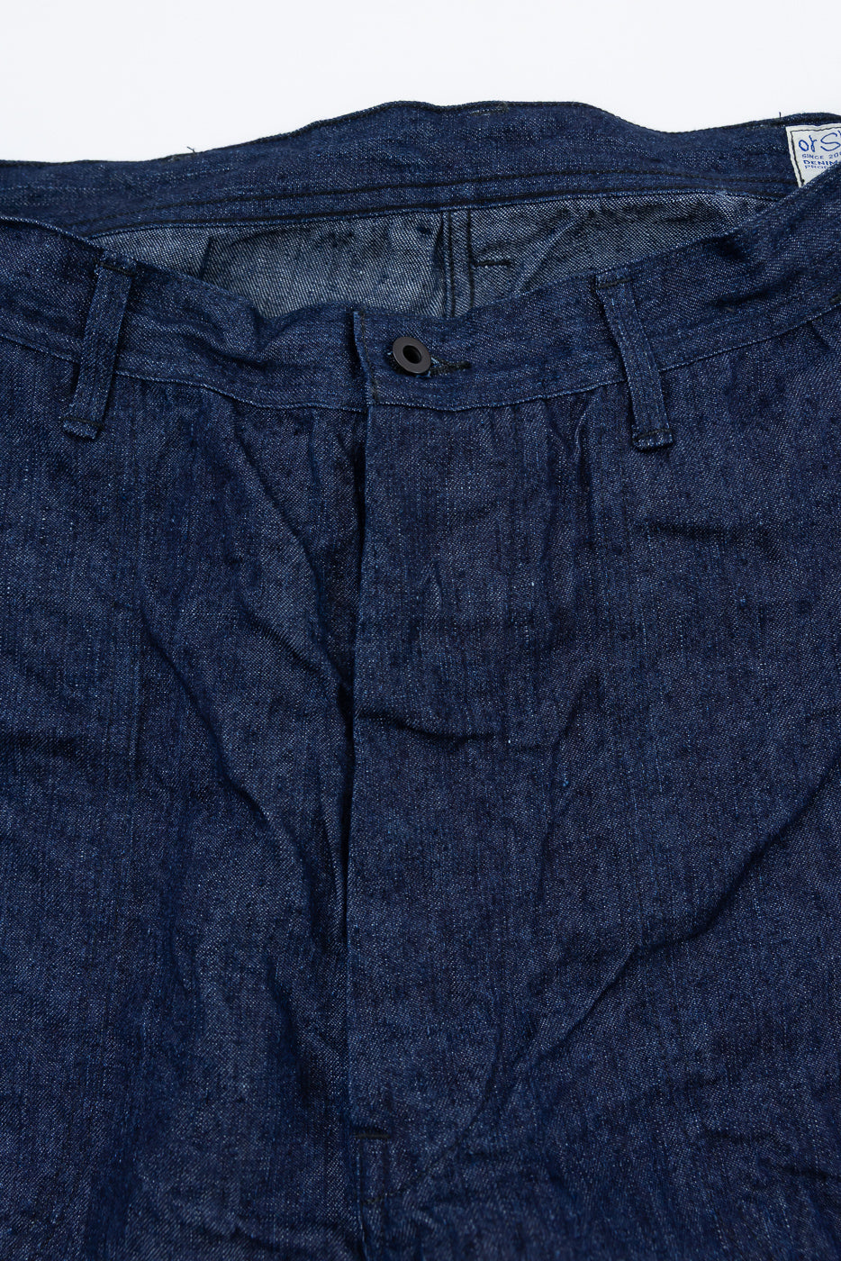 Close-up front view of dark denim pants showing button, waistband, and tag details