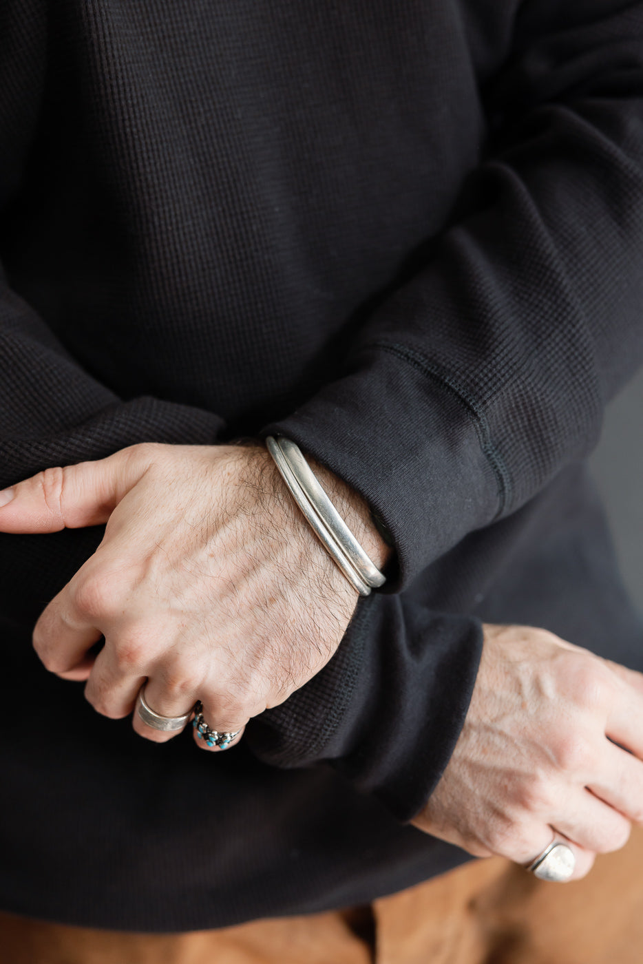 Close-up of a male model's wrists and hands wearing silver rings and bracelets over a black waffle knit long sleeve shirt.
