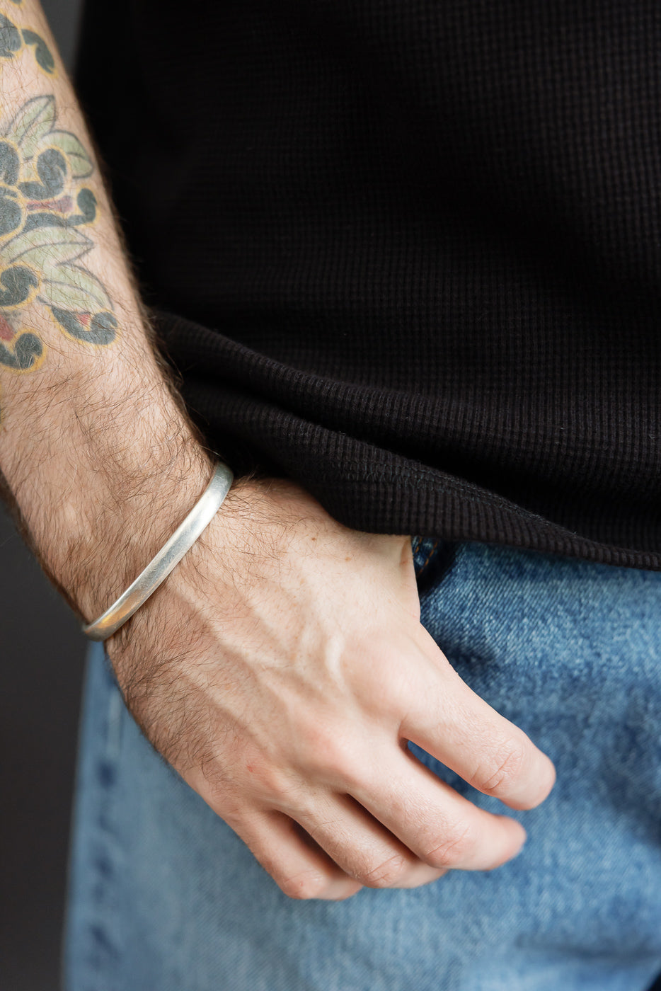 Close-up of a male model's wrist wearing a silver cuff bracelet, showing arm tattoo and black ribbed shirt.