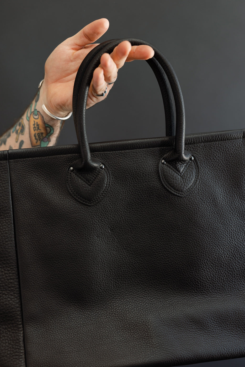 Close-up of black pebbled leather tote bag front, showing top handles, V-stitch detailing, and hand holding handle