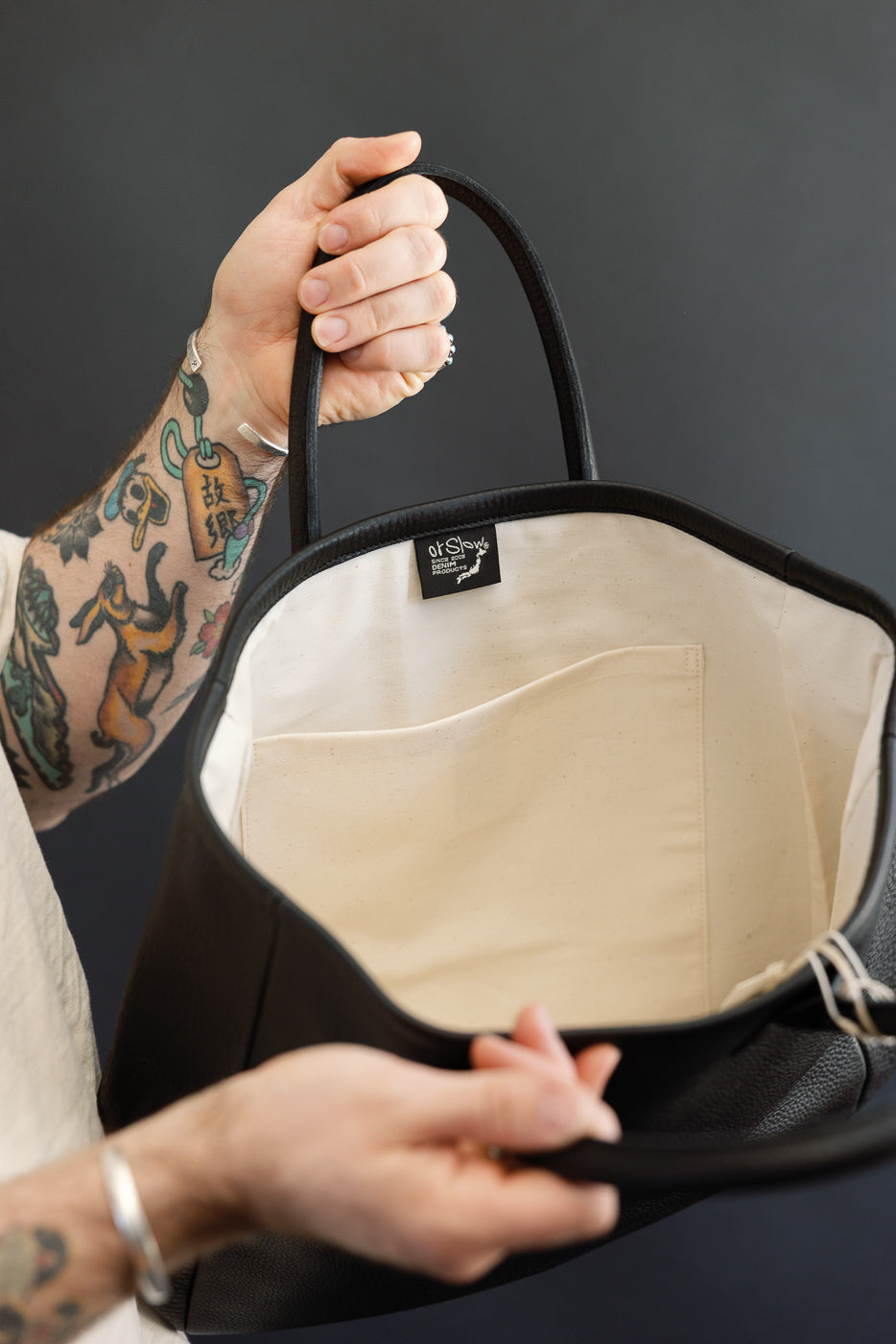 Close-up of black leather tote interior, showing cream lining, inner pocket, and 'atSlow' tag, held by tattooed male hand.