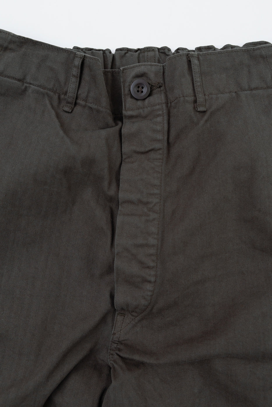 Close-up front view of the waistband and button closure on dark olive trousers.