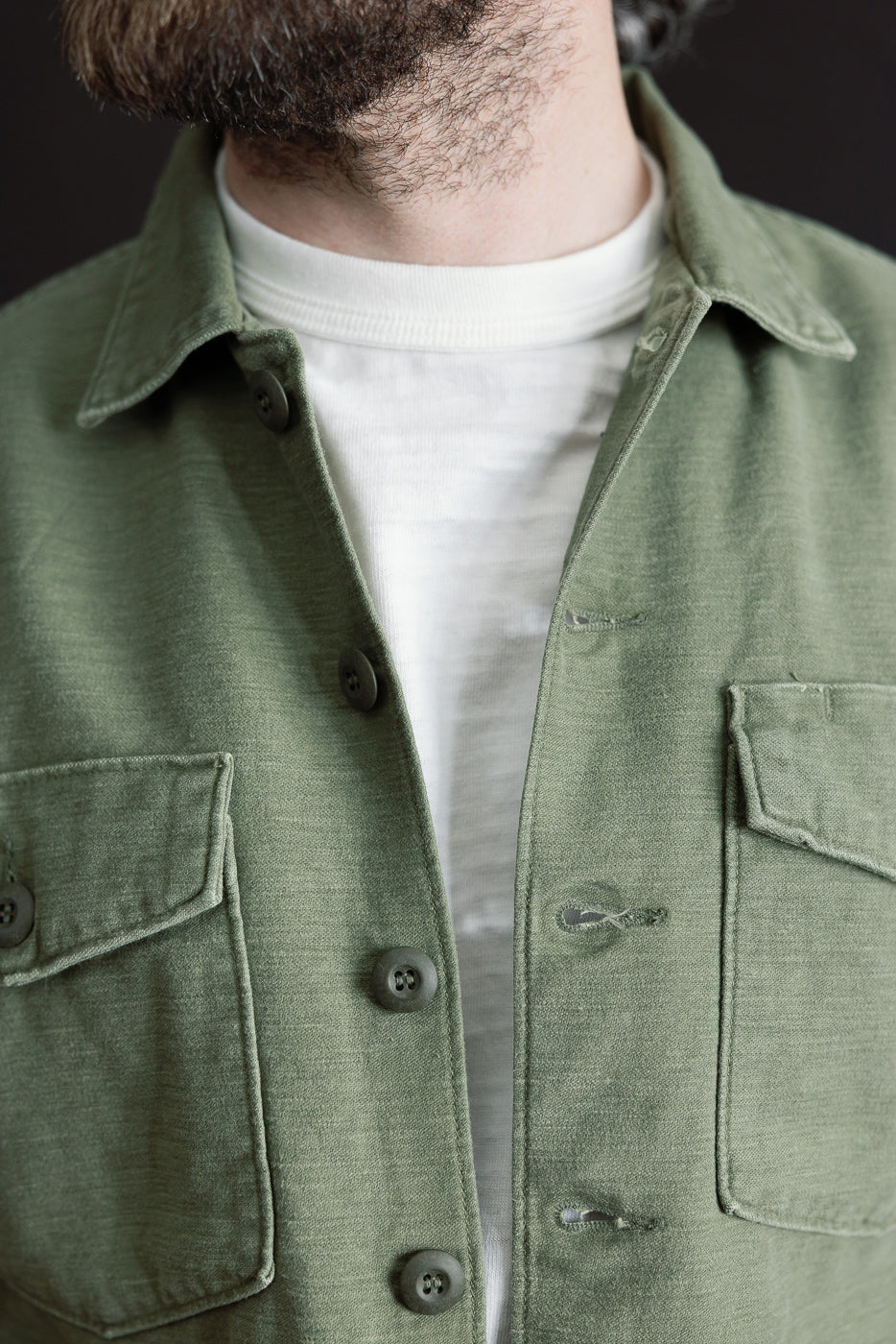 Close-up front view of orSlow 4-pocket fatigue shirt in dark olive, reverse sateen fabric, showing male model with athletic build, button details, collar, and chest pockets.
