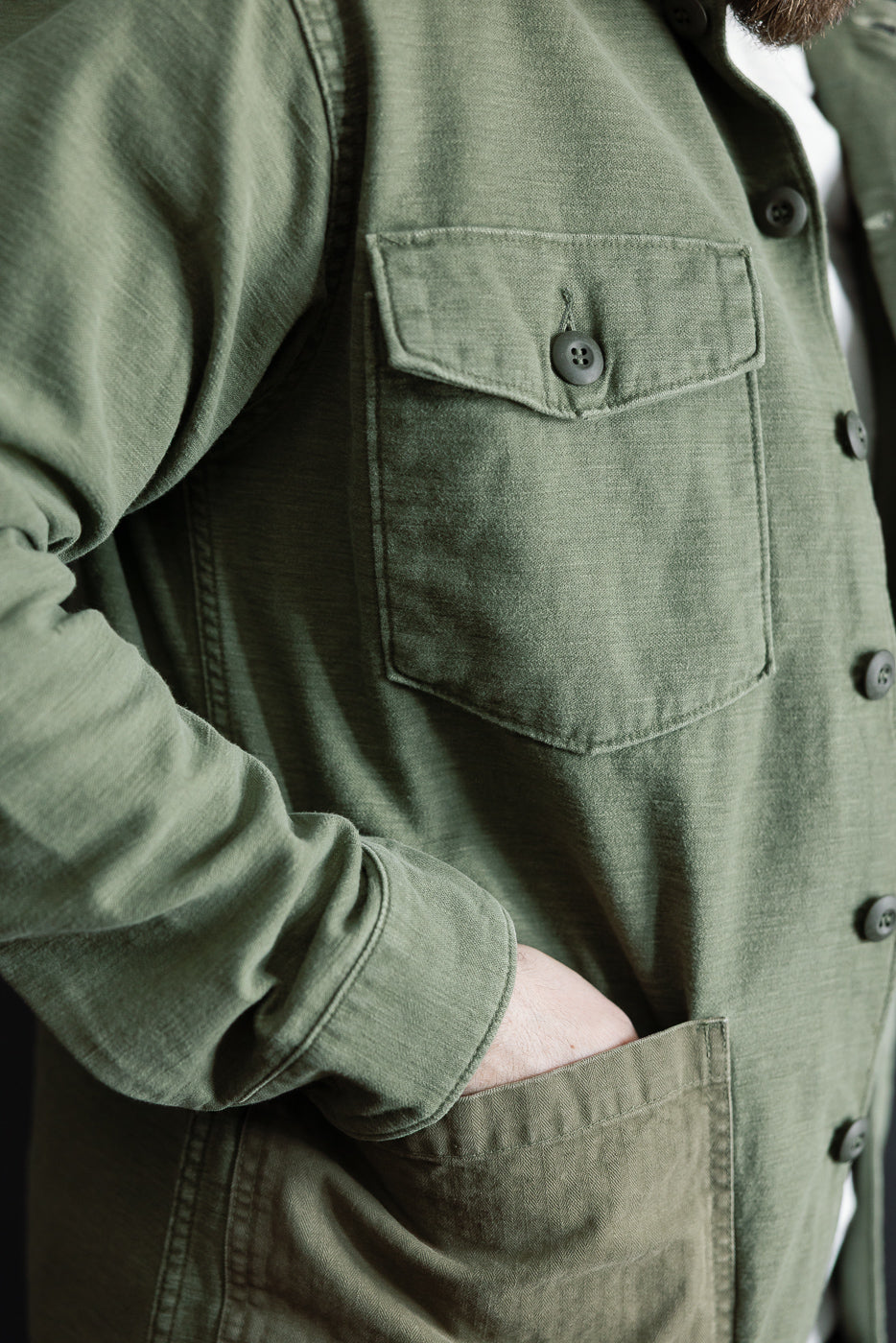 Close-up of orSlow dark olive fatigue shirt front left side, showing chest pocket, button detail, fabric texture on male model, 5'9