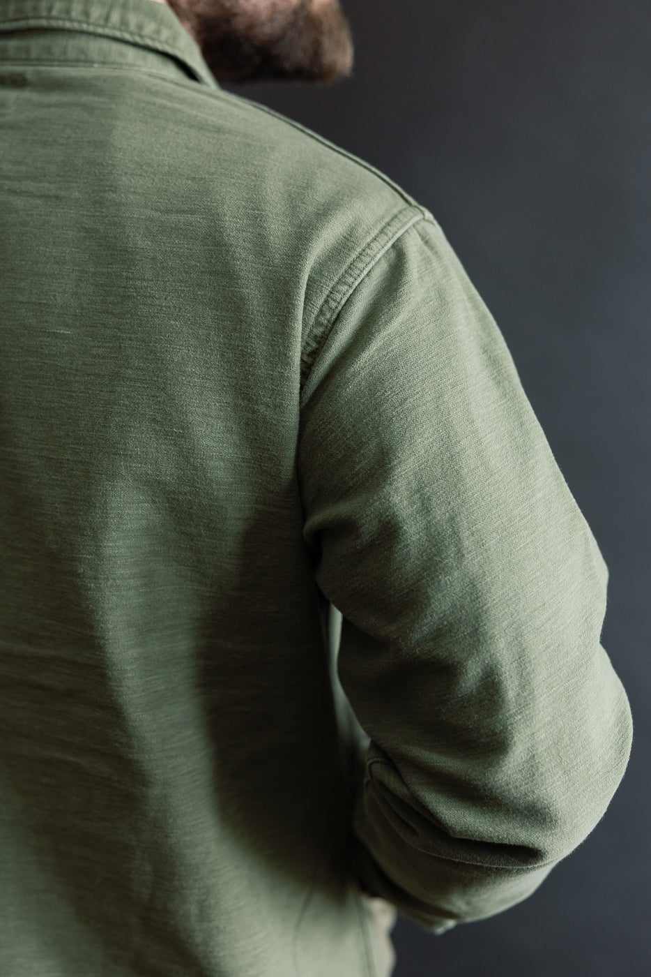 Close-up of male model's shoulder and upper back wearing dark olive fatigue shirt, rear view