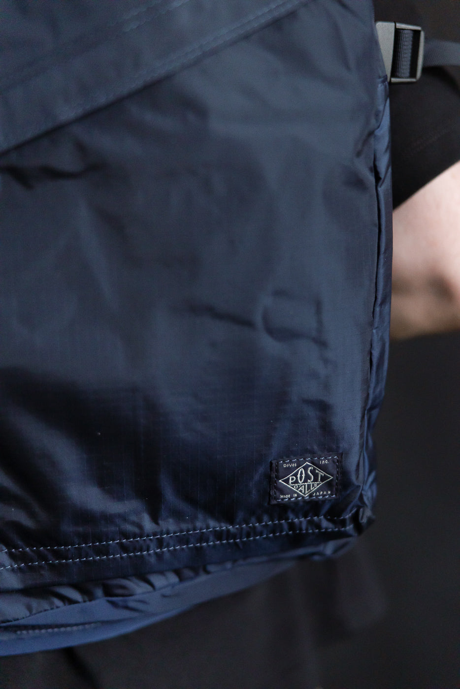 Close-up of male model wearing Post O’Alls navy nylon ripstop pack, front hem and logo tag detail