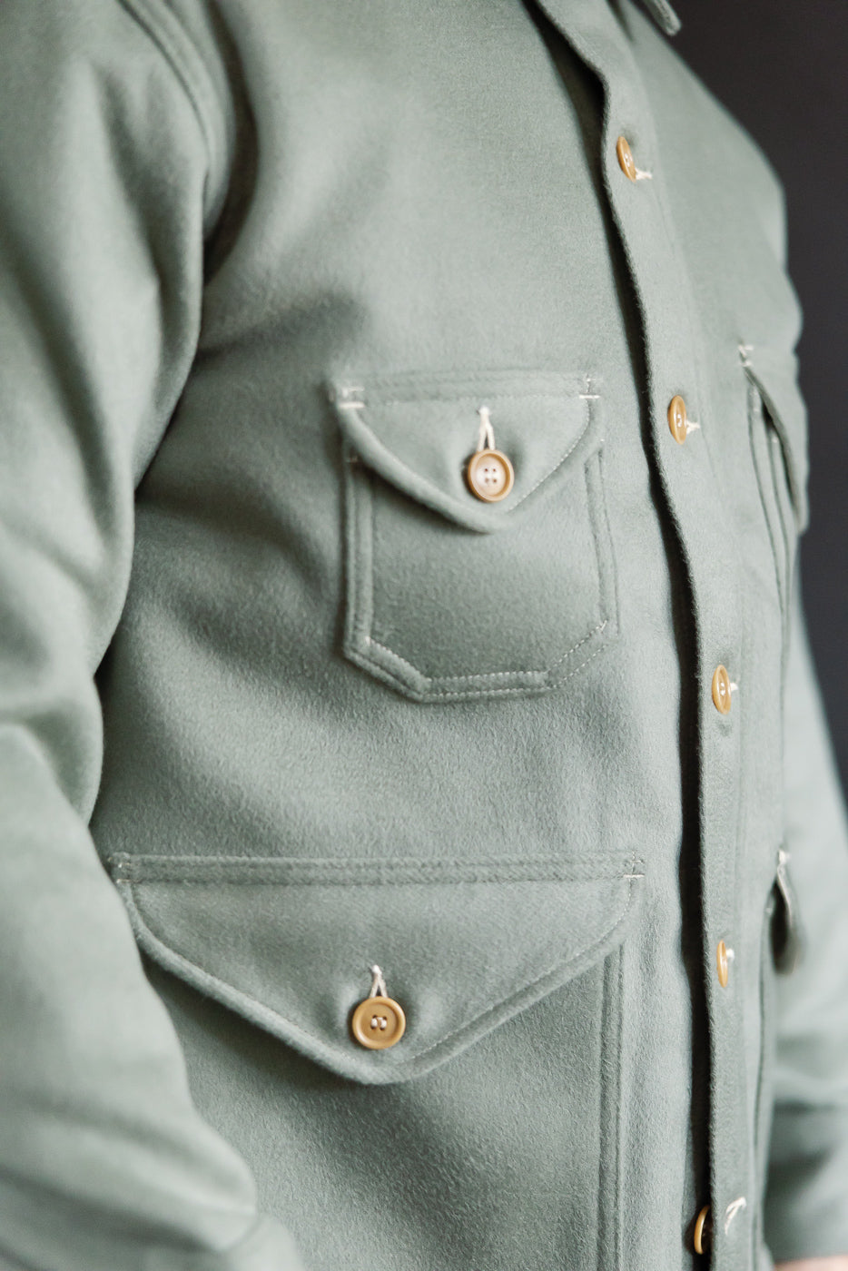 Close-up of male model's left front side wearing pale green wool jacket; shows buttoned flap pockets, tan buttons, and fabric texture. Model is 5'9