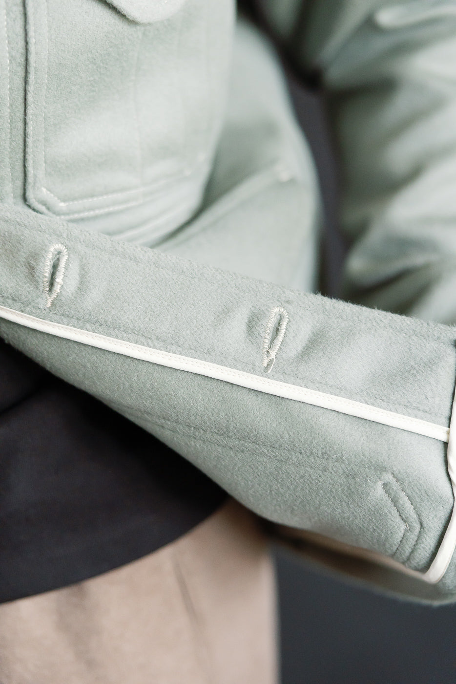 Close-up of pale green melton jacket sleeve cuff, showing buttonholes, contrast piping, and fabric texture, Dant contemporary casual outerwear.