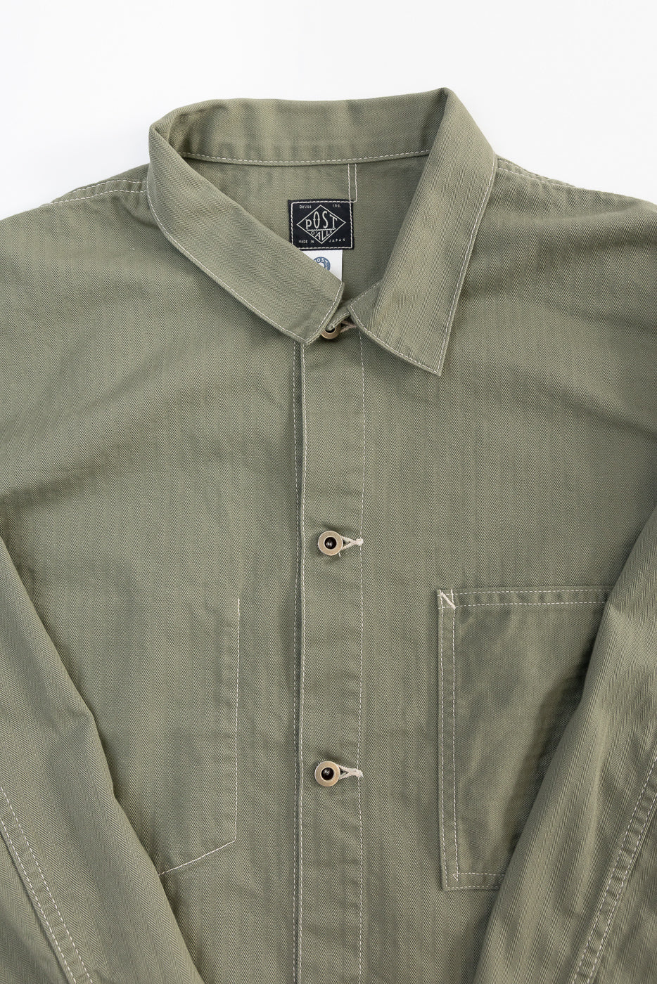 Close-up of front chest and collar on sage green utility jacket, showing buttons, pocket, and herringbone fabric texture.