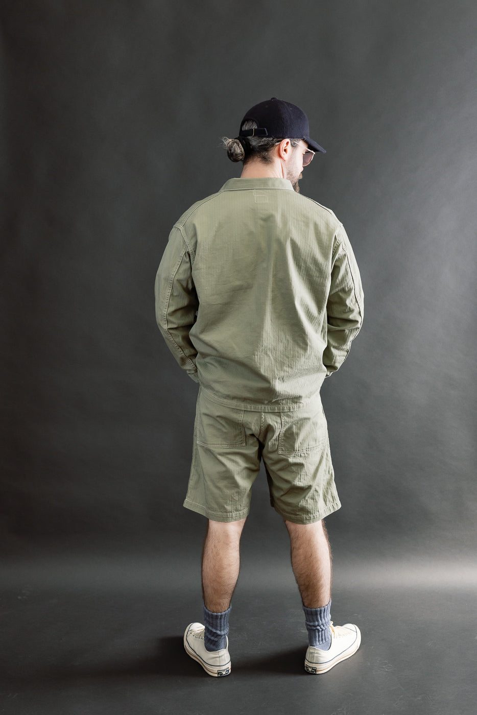Male model 5'9, 165lbs athletic build, full-body rear view in sage green 1930s utility jacket and shorts