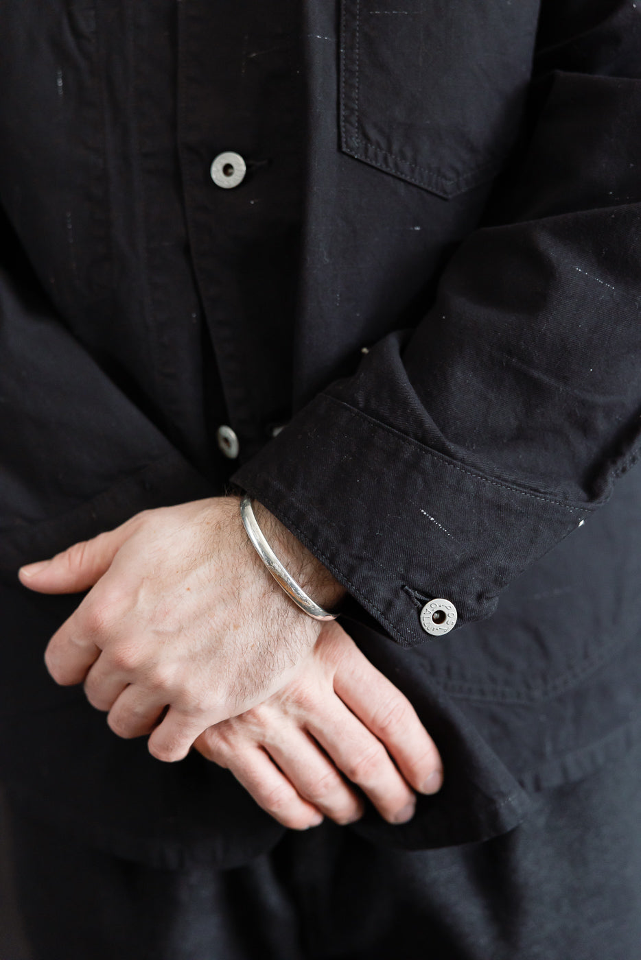 Close-up of front left cuff and chest pocket of black nep twill coverall on male model, showing button and fabric texture. Model is 5'9