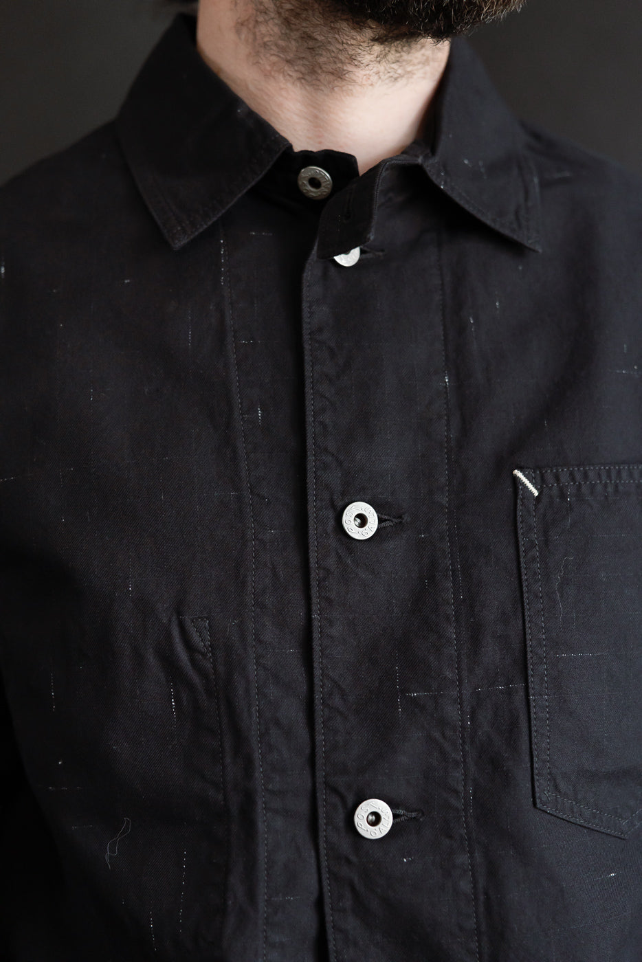 Close-up of male model's upper torso wearing black nep twill coverall, showing front buttons, collar, left chest pocket, and textured fabric. Model is 5'9