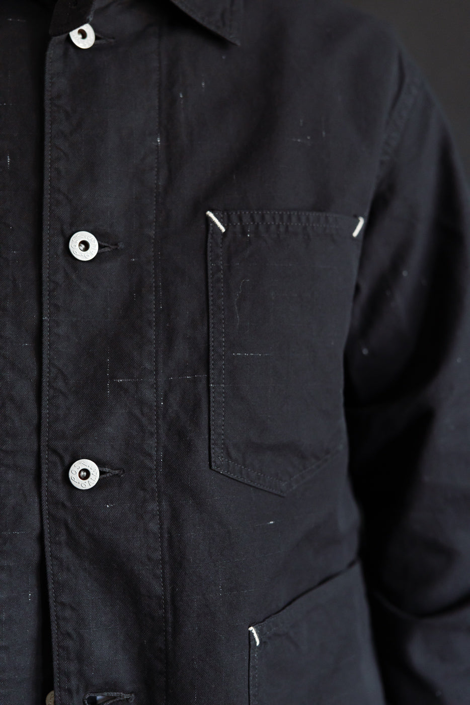 Close-up front of black Post 46 coverall jacket, showing chest pocket, metal buttons, and twill texture