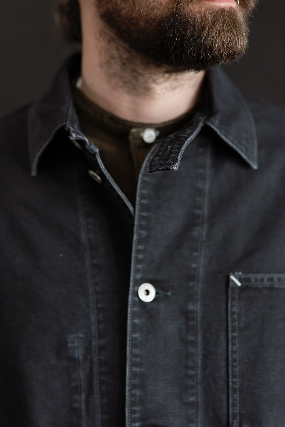 Close-up front view of washed black pigment dyed canvas coverall on male model, showing collar, white button, and chest pocket. Model is 5'9