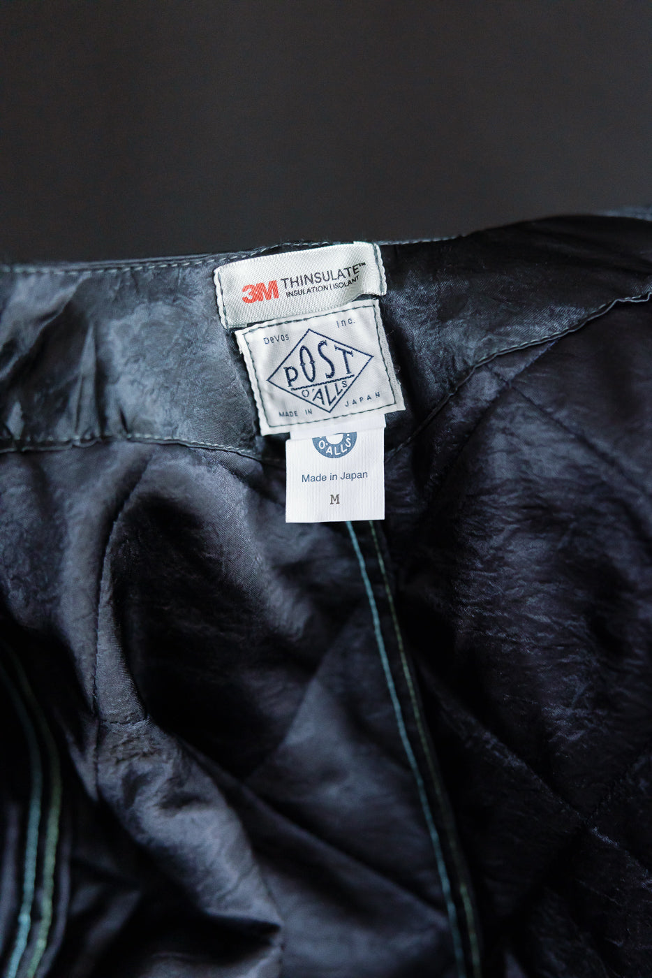 Close-up of Post O'Alls quilted acetate sateen coverall interior neck tags, showing 3M Thinsulate, Made in Japan label, and inside fabric texture, dark grey.