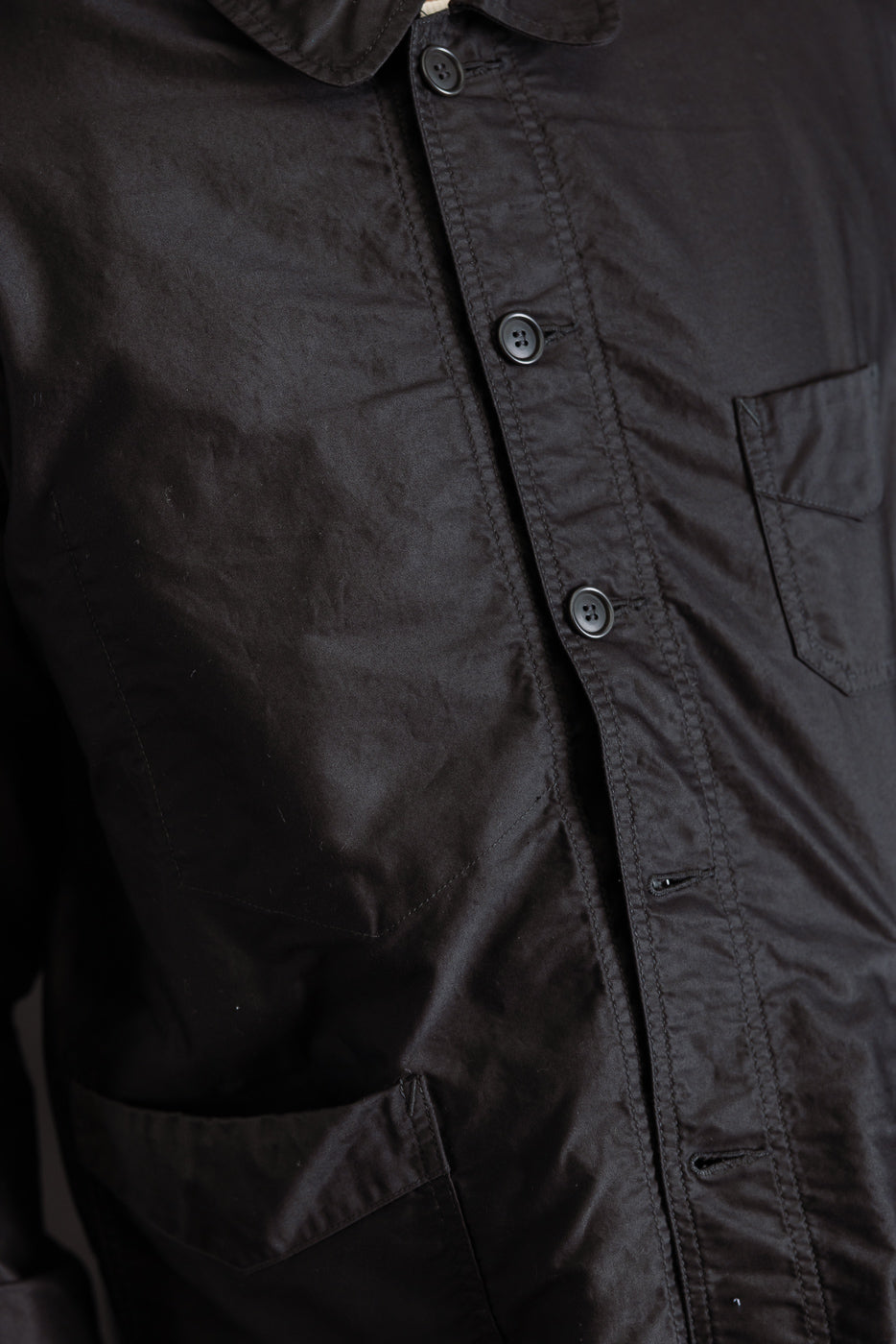 Close-up of front left side of black moleskin work jacket, showing buttons, collar, chest and waist pockets, fabric texture.