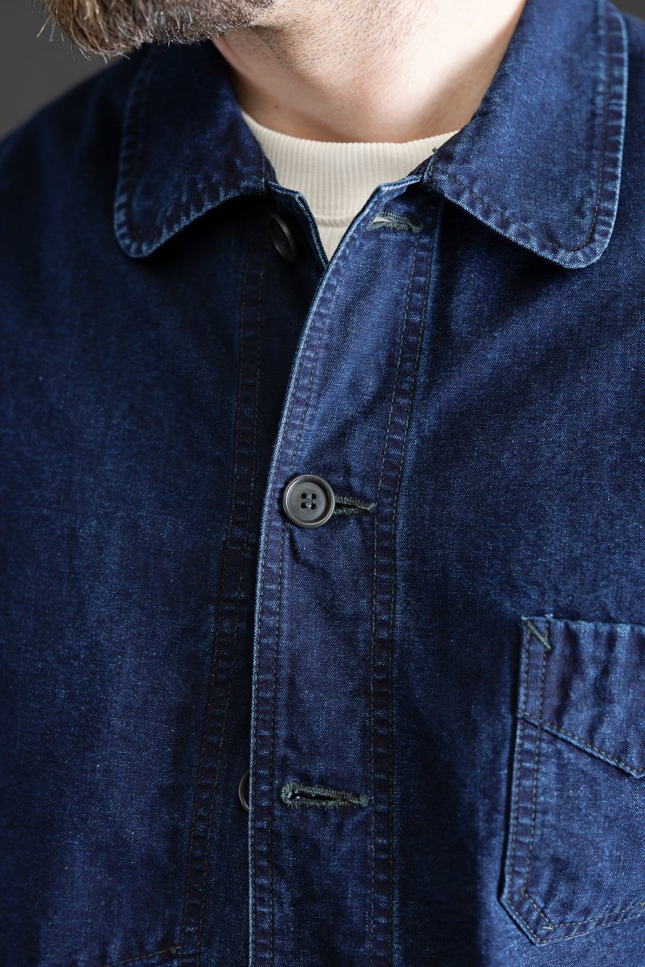 Close-up front view of deep washed indigo canvas work jacket on male model, showing buttons, collar, pocket, and fabric texture, model is 5'9