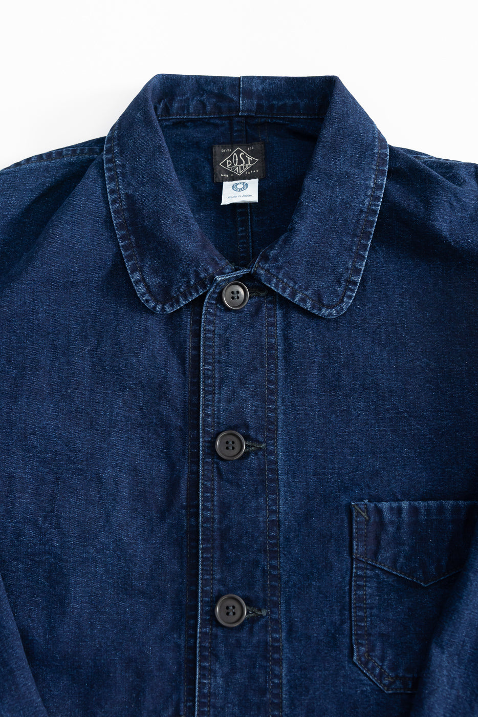 Close-up front of indigo washed canvas chore jacket, showing buttons, collar, left chest pocket, and visible tags.