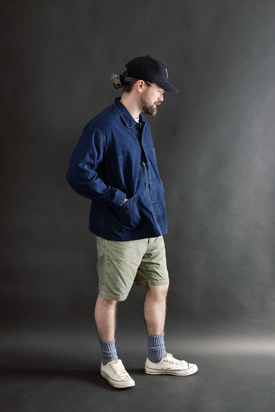Full body side view of male model, 5'9, 165lbs athletic build, wearing deep washed indigo canvas work jacket with visible front buttons and patch pockets, paired with green shorts and white sneakers.