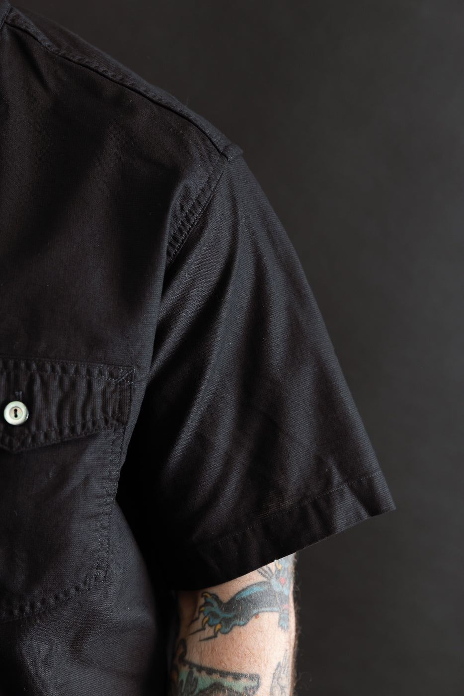 Close-up of men's short sleeve black shirt, left chest pocket, button, and sleeve fabric details, worn by male model, 5'9