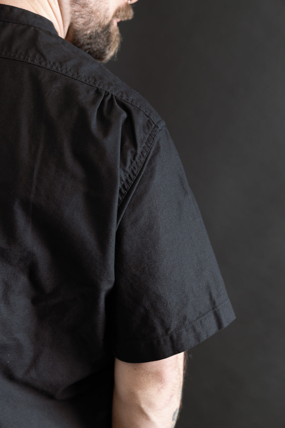 Close-up of male model's upper back and shoulder wearing black short sleeve shirt, left side view. Model is 5'9
