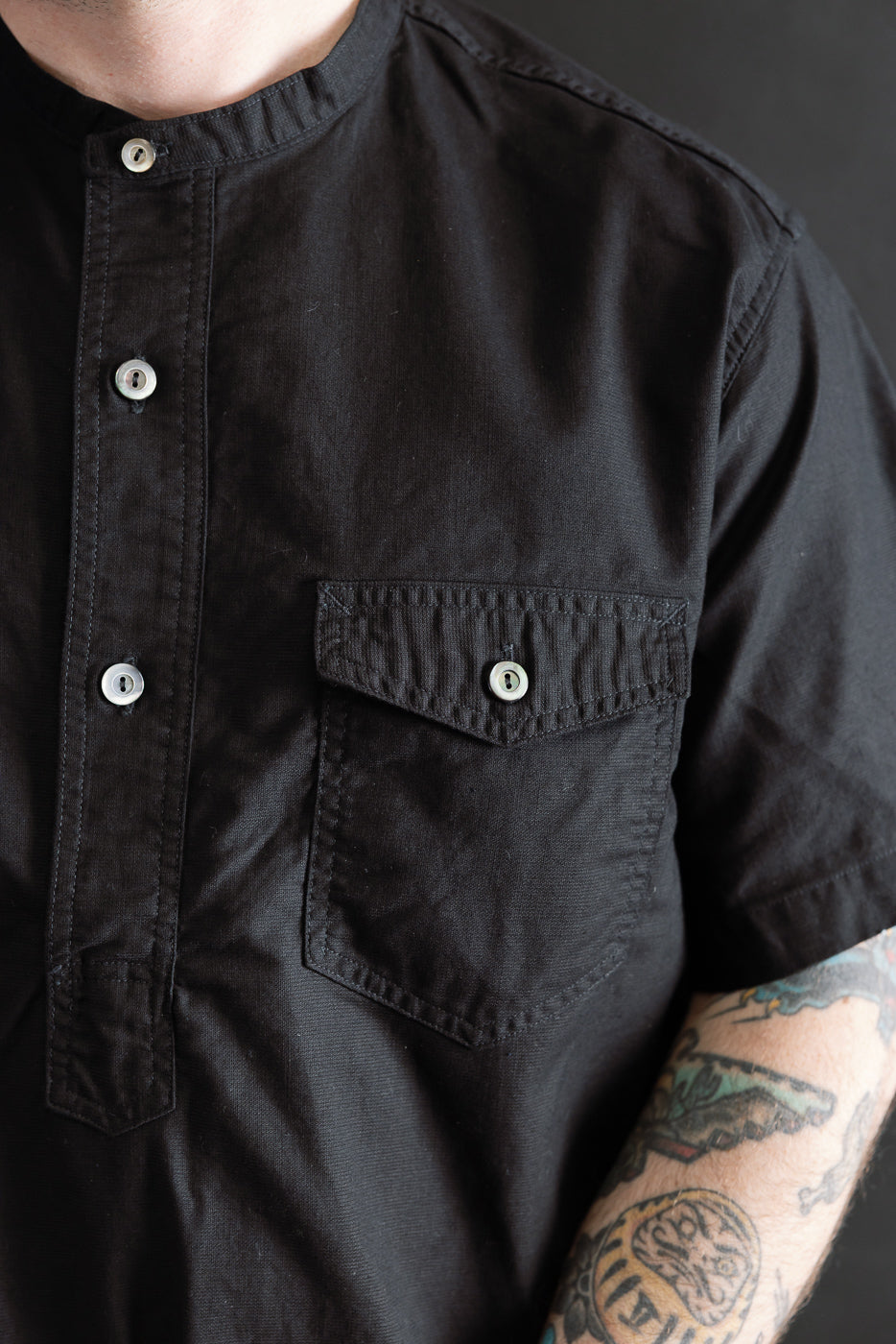 Close-up front view of black short sleeve band collar shirt, chest pocket, worn by male model 5'9