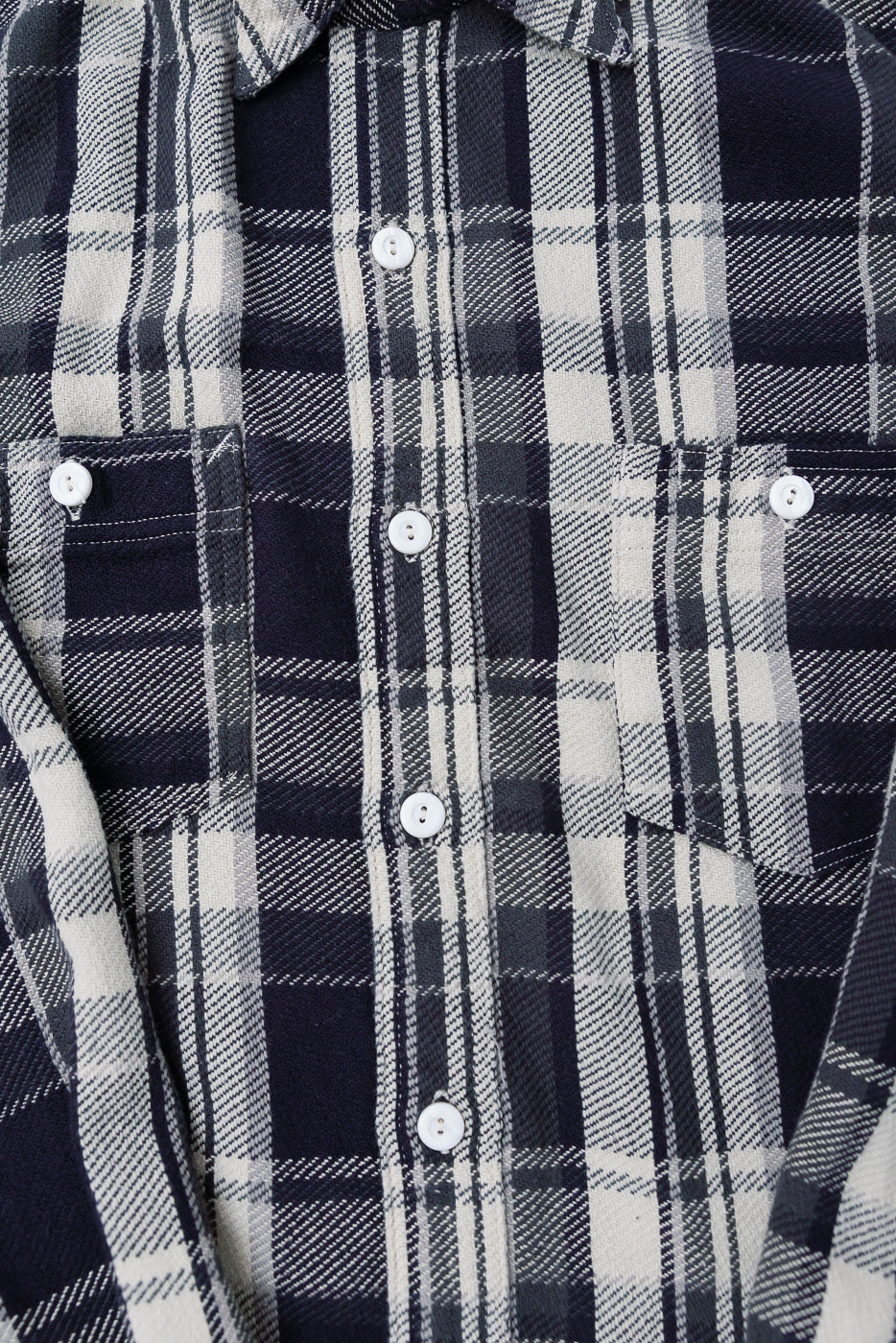 Close-up of front chest and button placket of grey plaid cotton flannel shirt, showing fabric texture, white buttons, and dual front pockets.