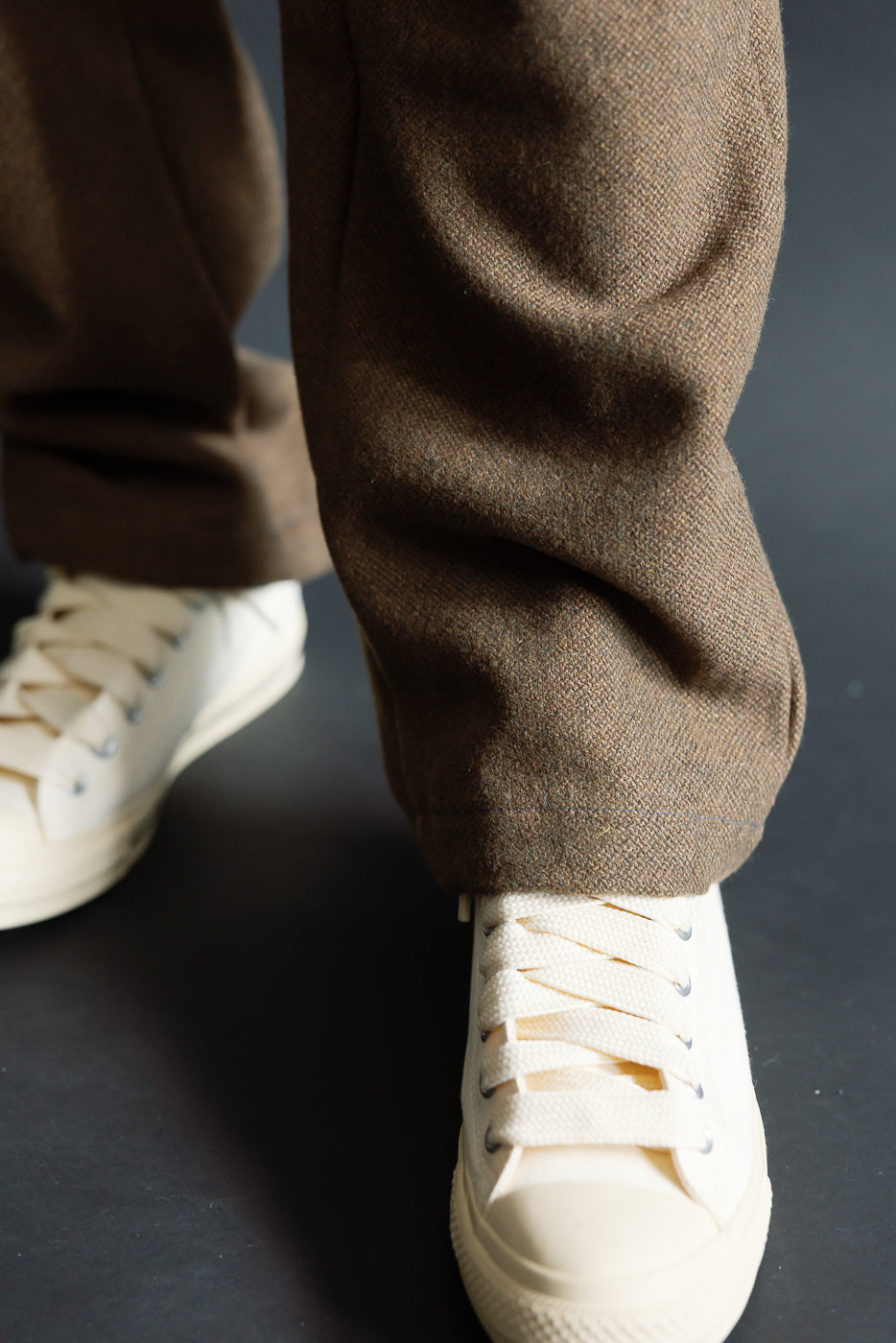Close-up of male model's legs wearing brown tweed pants and white sneakers, hem detail visible, front view. Model is 5'9