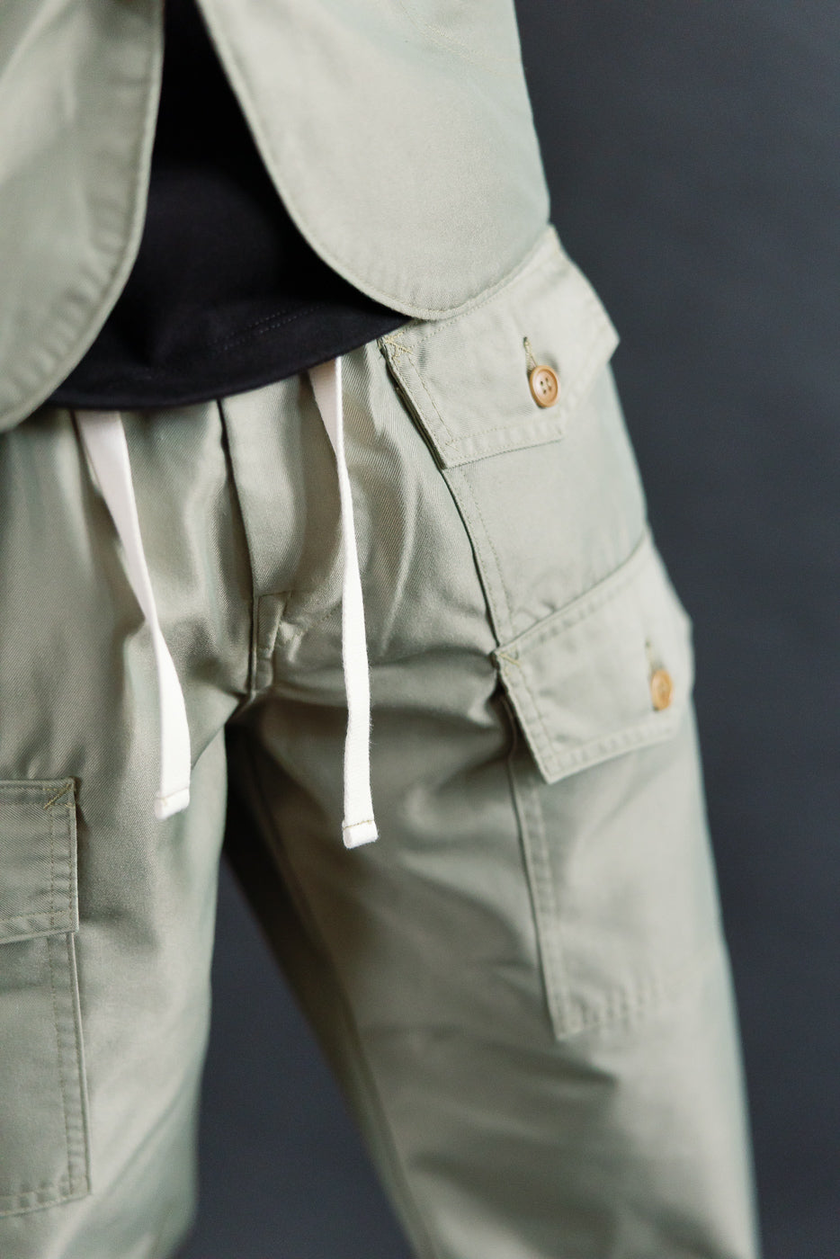 Close-up front view of Post O'Alls khaki French twill pants on male model, showing drawstring waist and cargo pocket button details. Model is 5'9