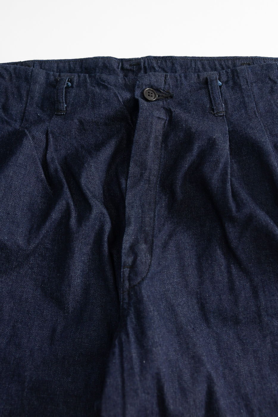 Close-up of front waist and button details on dark indigo denim pants, showcasing fabric texture.