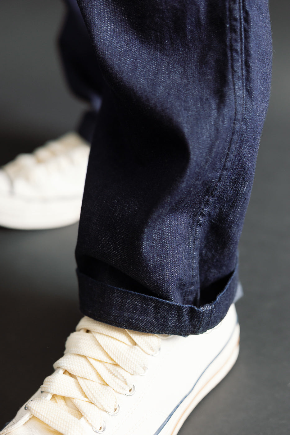 Close-up of dark indigo denim pant leg hem and white sneaker, side view of cuff detail, on-body male model.