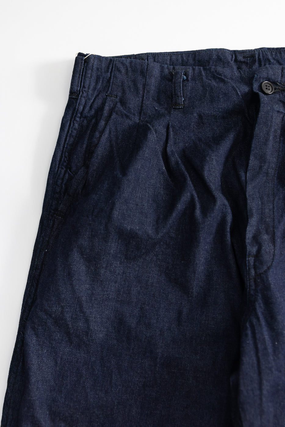 Close-up of front left side of dark indigo denim pants showing waistband, pleats, and button detail
