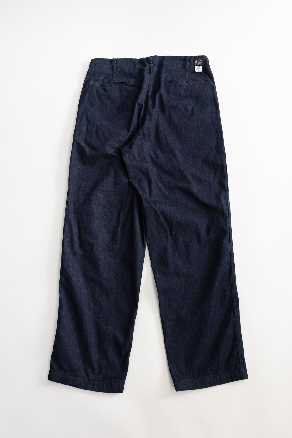 Flat lay of dark indigo denim pants, back view, showing rear pockets and waistband tag detail