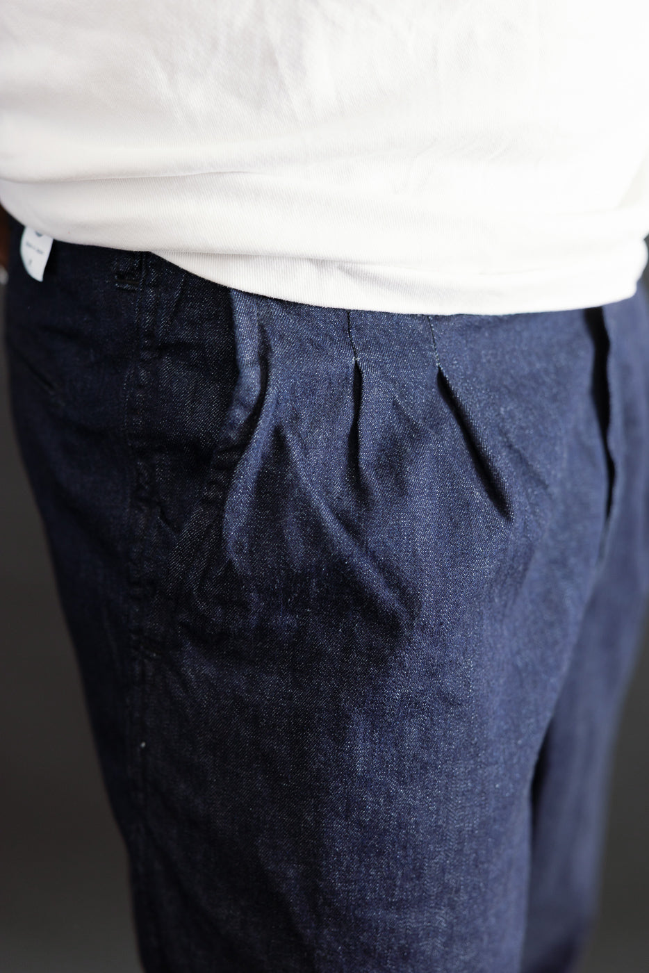 Close-up of front left side of 8oz dark indigo denim pants on male model, showing pleats and pocket. Model is 5'9