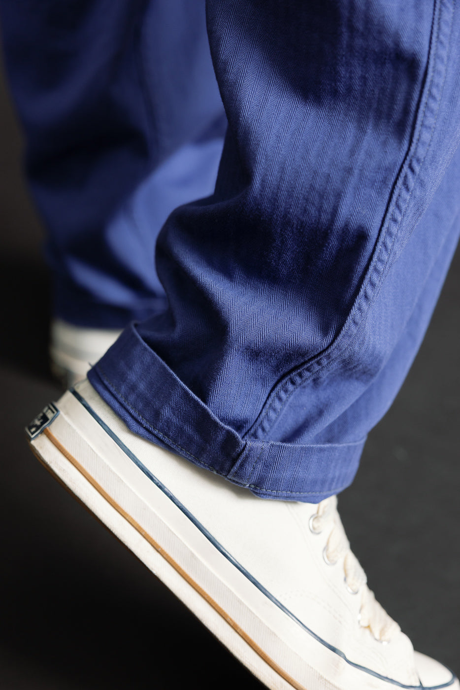Close-up of male model's lower leg wearing blue herringbone pants right side, showing hem stitching and fabric texture, model is 5'9