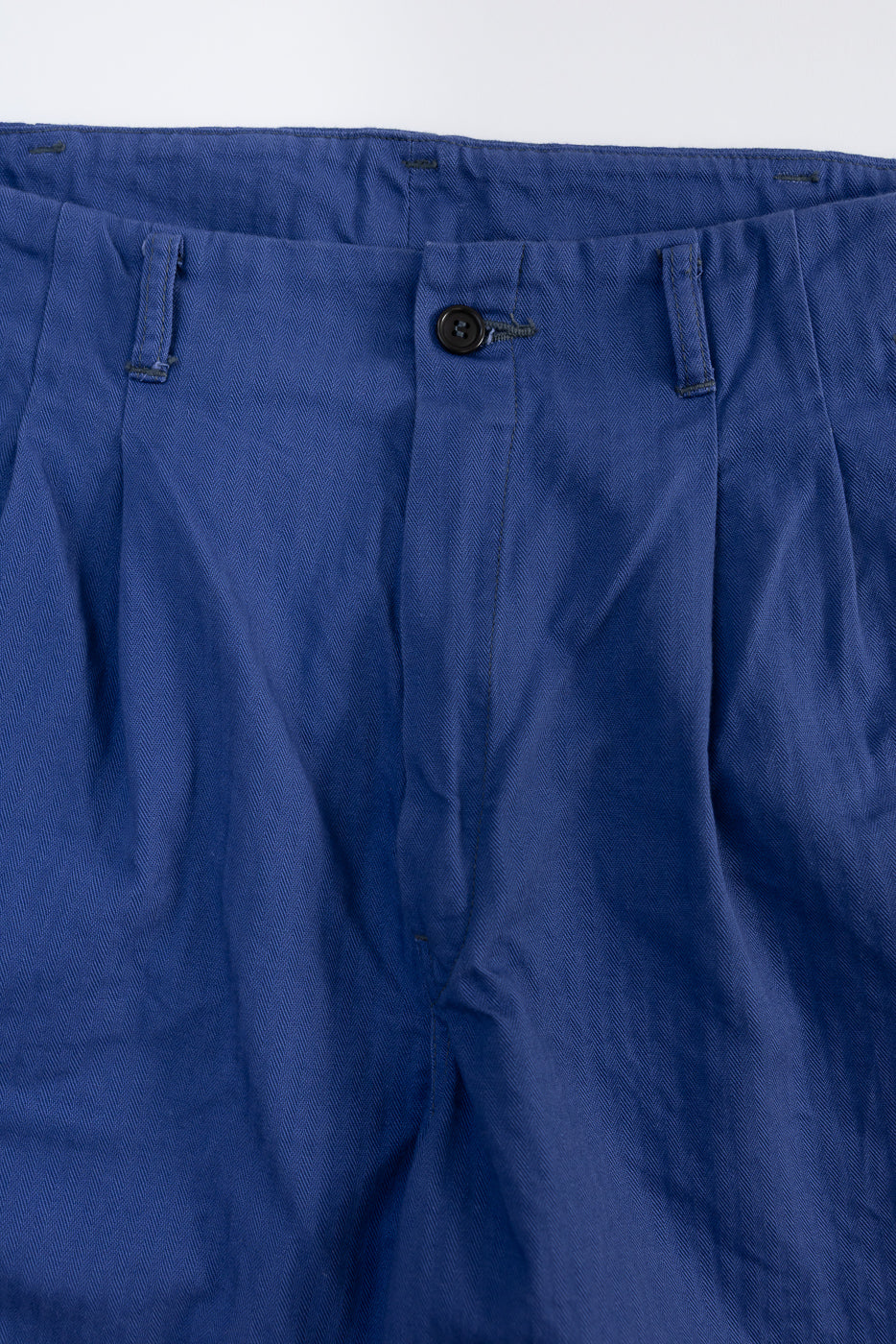 Close-up of front waistband and button fly on blue herringbone pants, showcasing fabric texture.