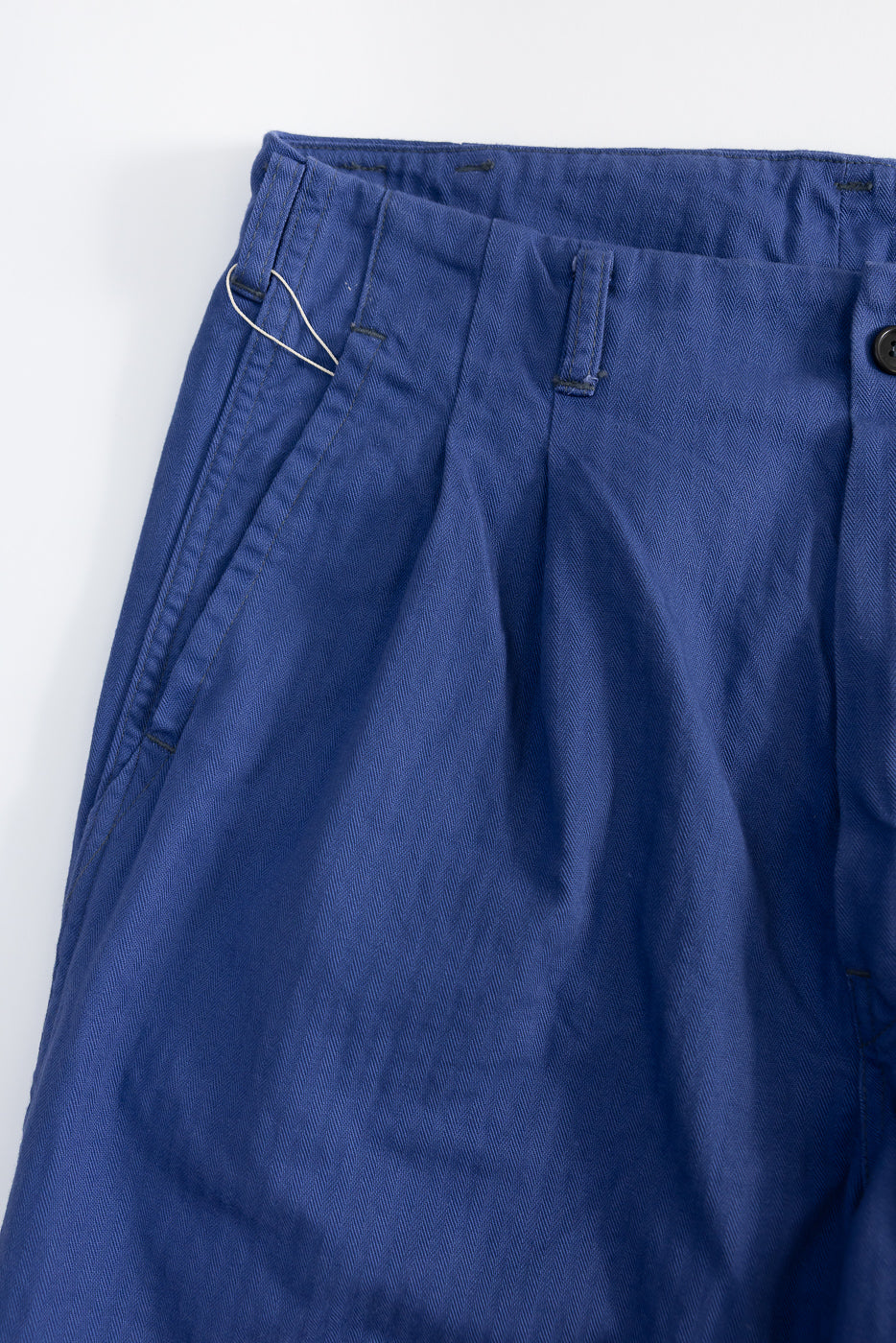 Close-up of front left side of blue herringbone twill pants, showing waistband, pleat, and pocket detail.