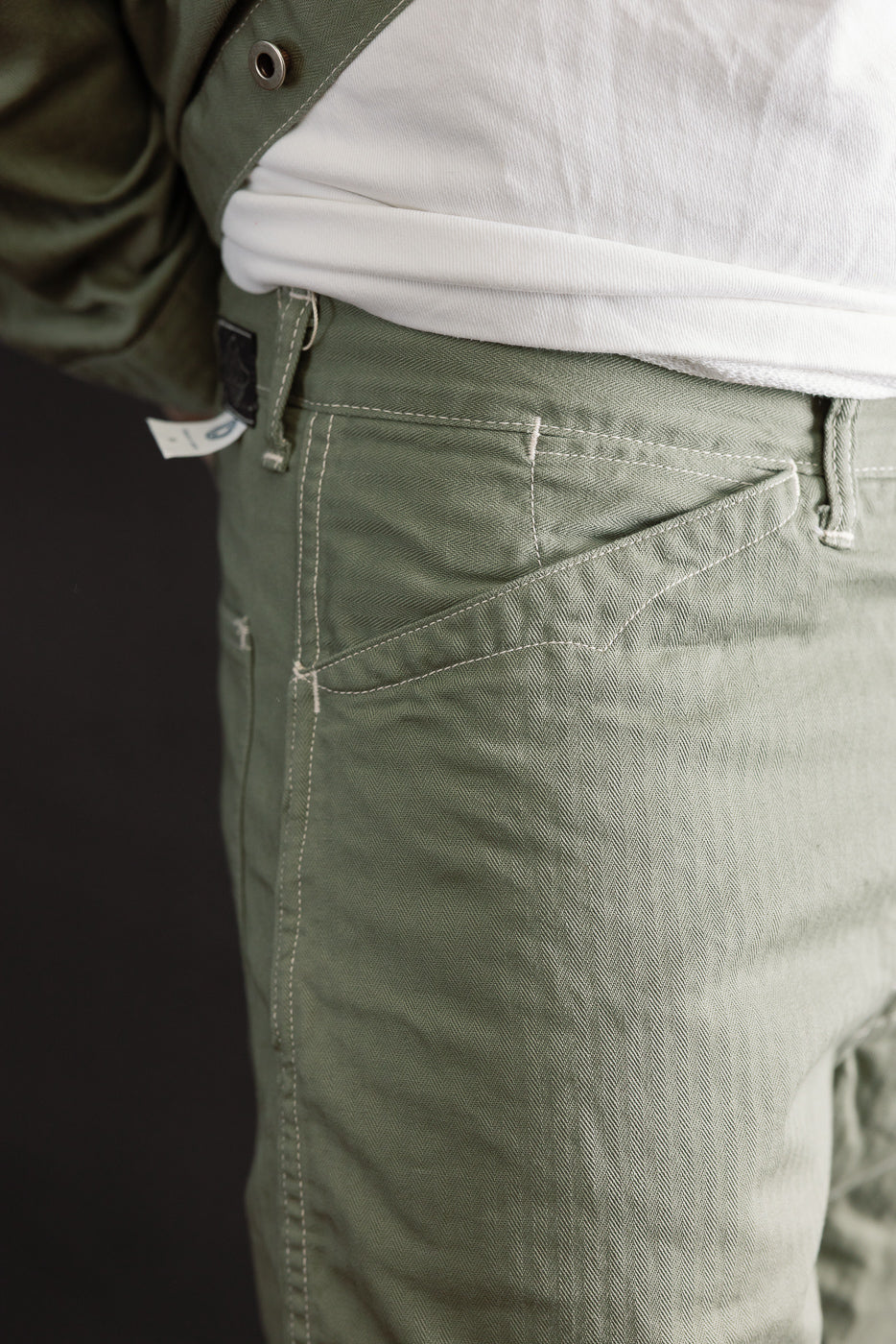 Close-up of right front pocket and waist of sage herringbone shorts on male model, 5'9, 165lbs athletic build.