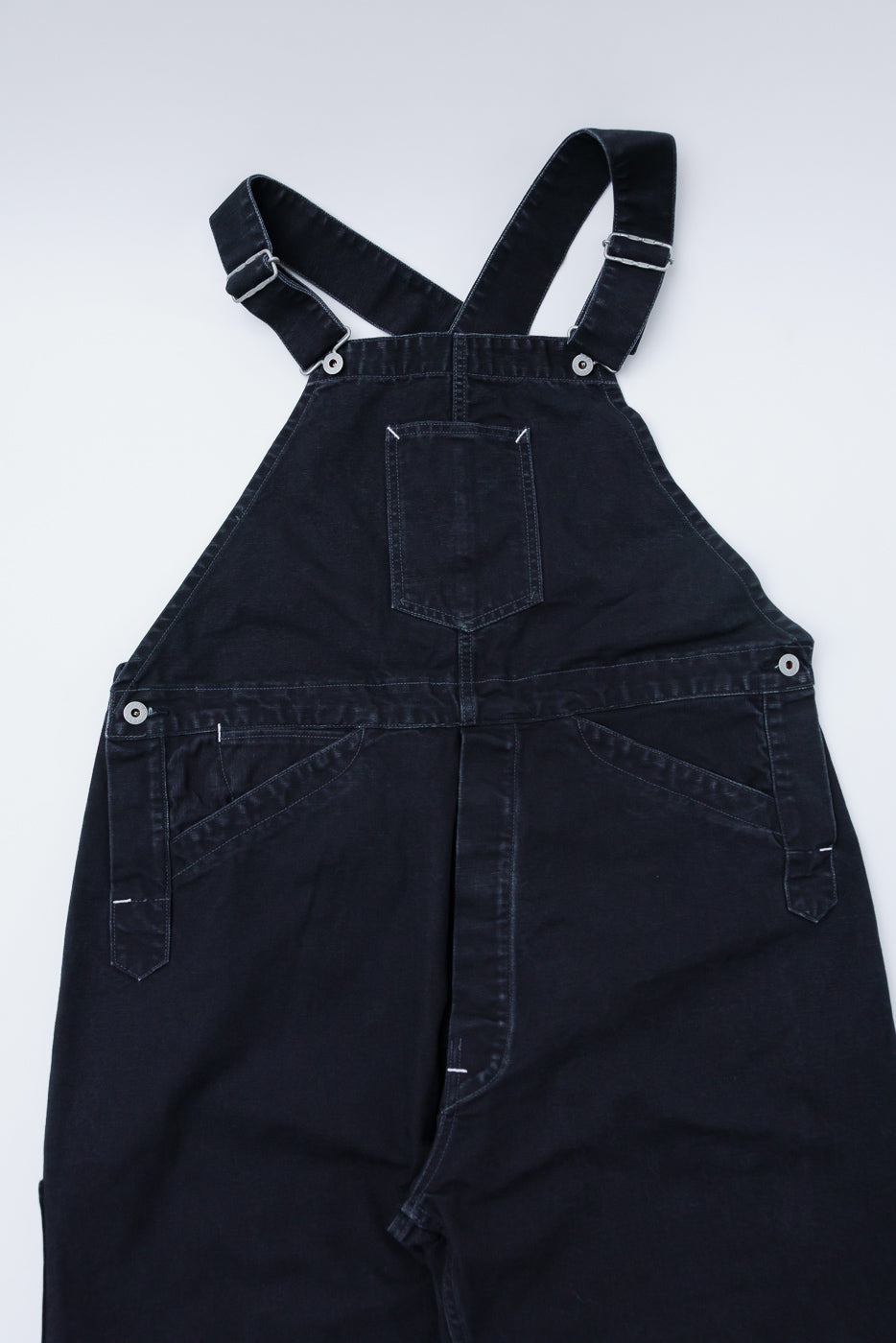 Front flat lay close-up of black pigment-dyed canvas overalls, featuring pocket and strap details, Dant contemporary apparel.