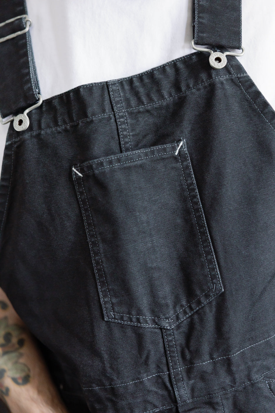 Close-up of front chest pocket and straps on black pigment-dyed canvas overalls, worn by male model, athletic build, Dant casual apparel.