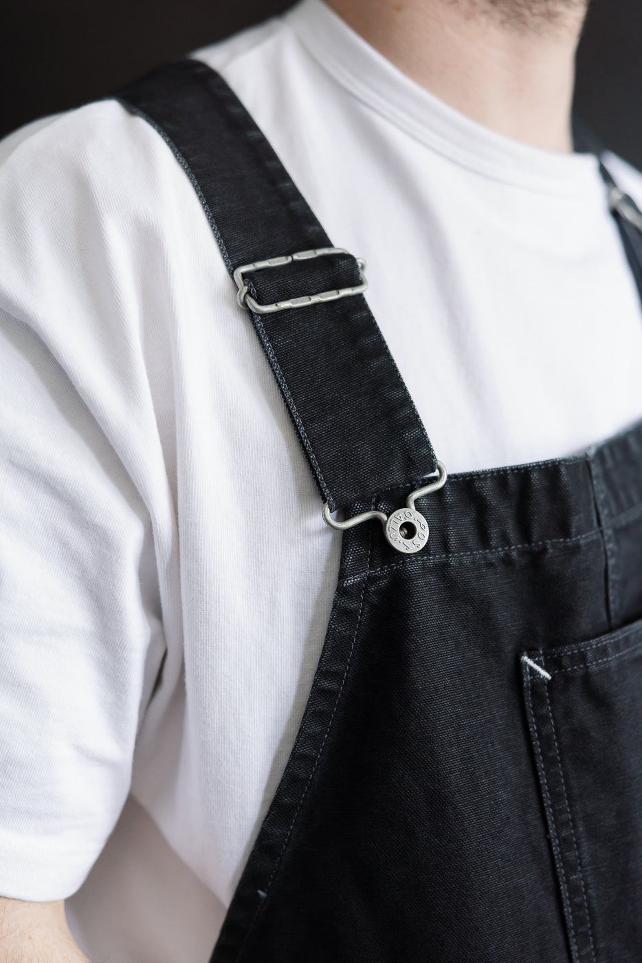 Close-up of male model, 5'9” 165lbs athletic build, wearing pigment dyed black canvas overalls front bib and strap detail from Dant
