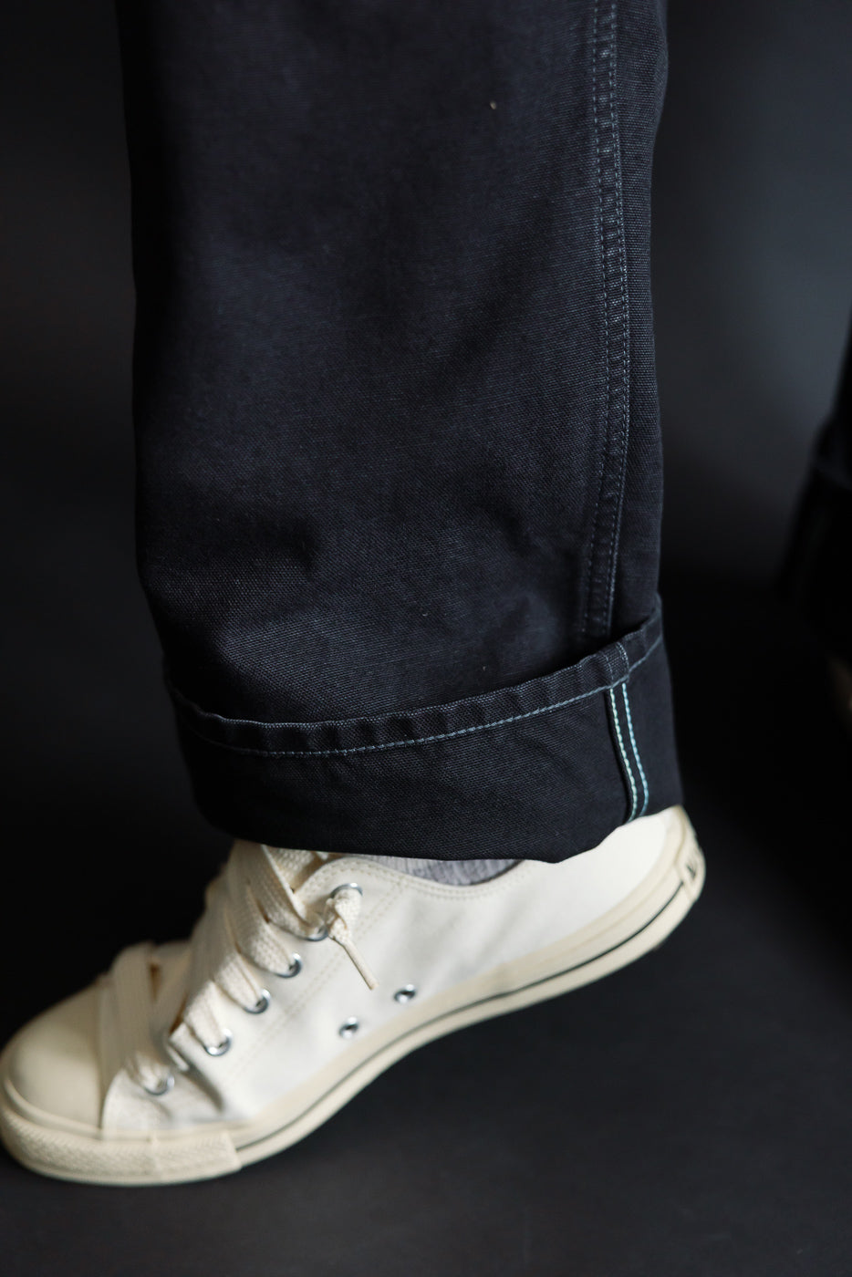 Close-up of black pigment dyed canvas overalls hem, side view, with cream canvas sneaker; male model, 5'9