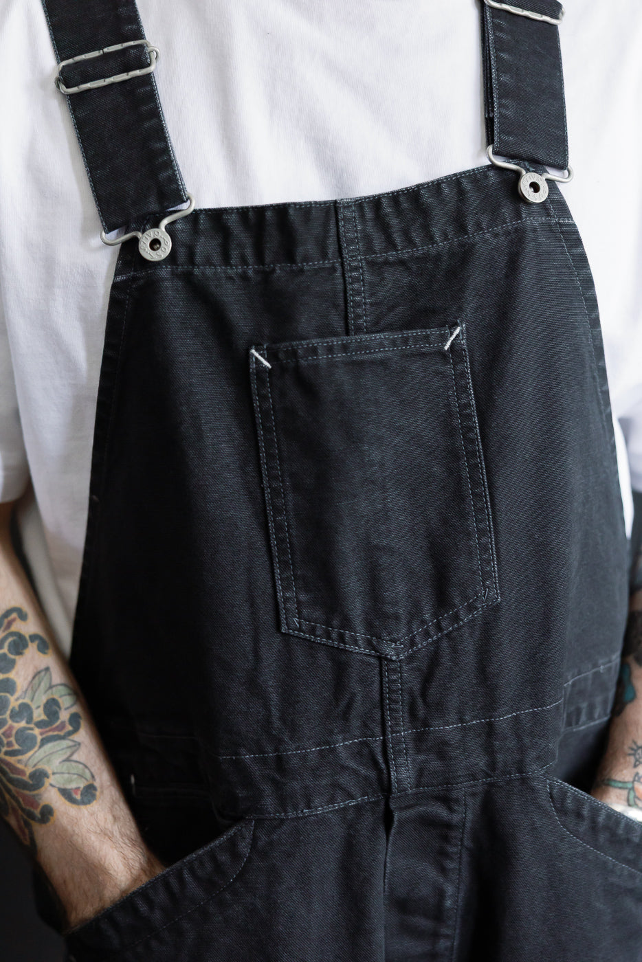 Close-up front view of black pigment-dyed canvas overalls on male model, showing chest pocket and strap details. Model is 5'9