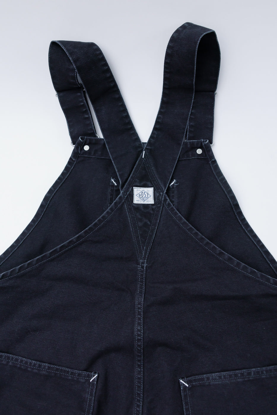 Close-up front view of Post Overalls 46 overalls in black pigment dyed canvas, showing pocket and strap detail, with visible stitching and brand tag.