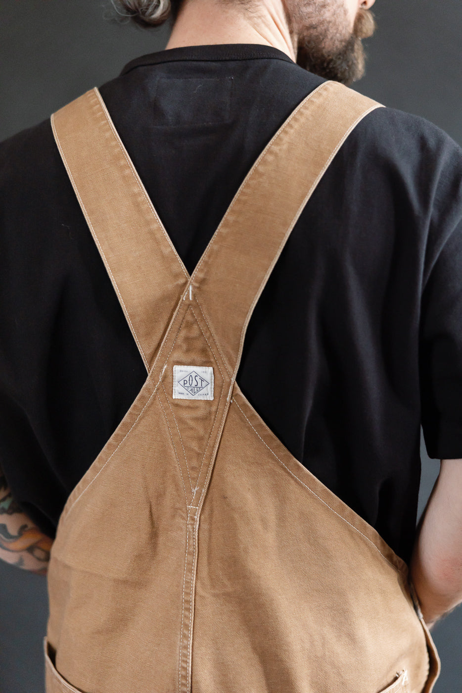 Close-up of men's washed brown pigment-dyed canvas overalls back, showing crossed straps and Post Overalls patch, on 5'9