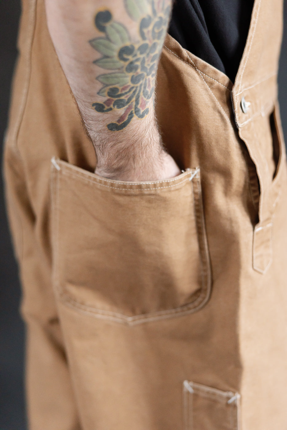 Close-up side view of men's brown pigment-dyed canvas overalls front pocket detail, model is male, 5'9
